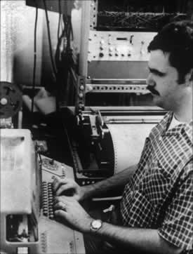Figure 1.     MIT Master's candidate, who is blind and a Braille reader, is using the prototype  MIT Braillemboss (in the background) while on a summer job. He types on a  standard teletypewriter, the signals go over telephone lines to MIT where  a computer with the DOTSYS translating program translates the copy to Braille,  which is then transmitted to the Braillemboss. 
